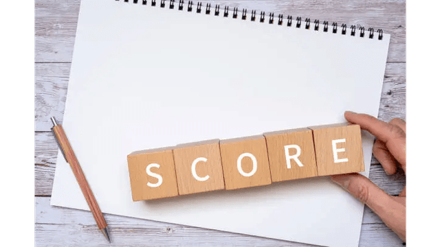 A hand placing wooden blocks spelling "SCORE" on a blank notebook with a pencil on a white wooden surface.