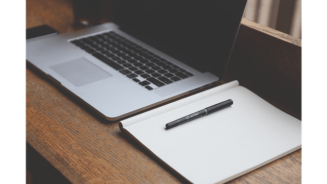 A laptop sits on a wooden desk next to a notepad and pen.